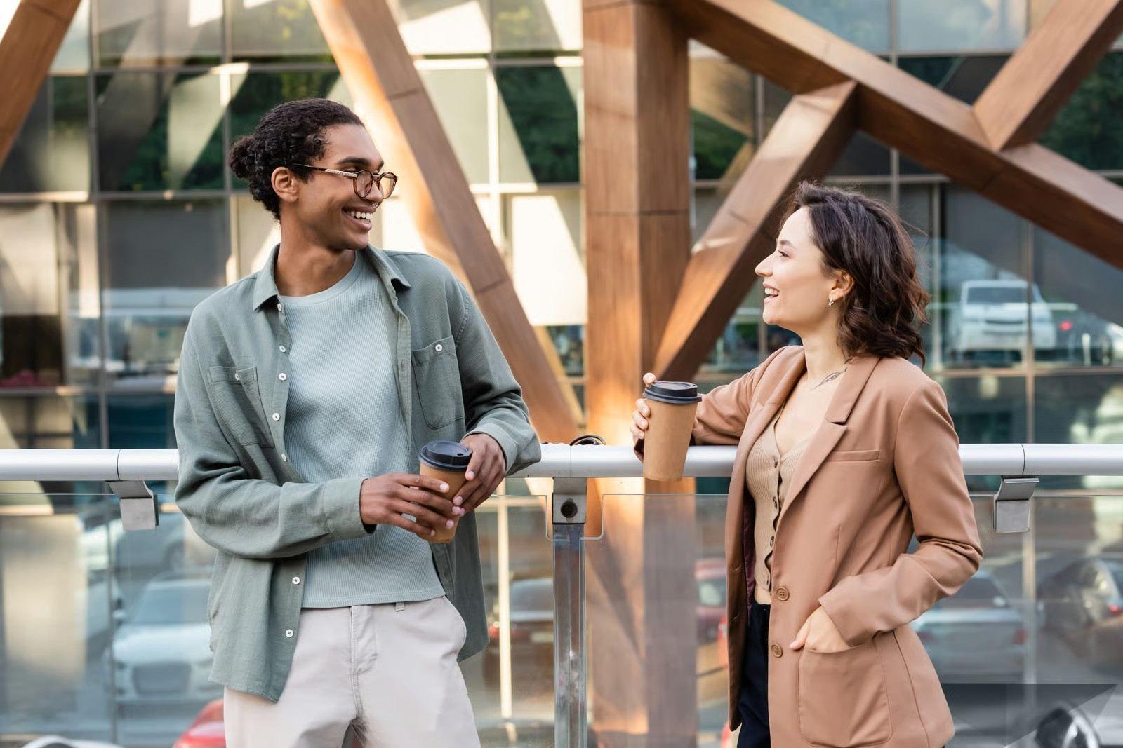 Two smiling professionals chatting outdoors while holding takeaway coffee cups.
