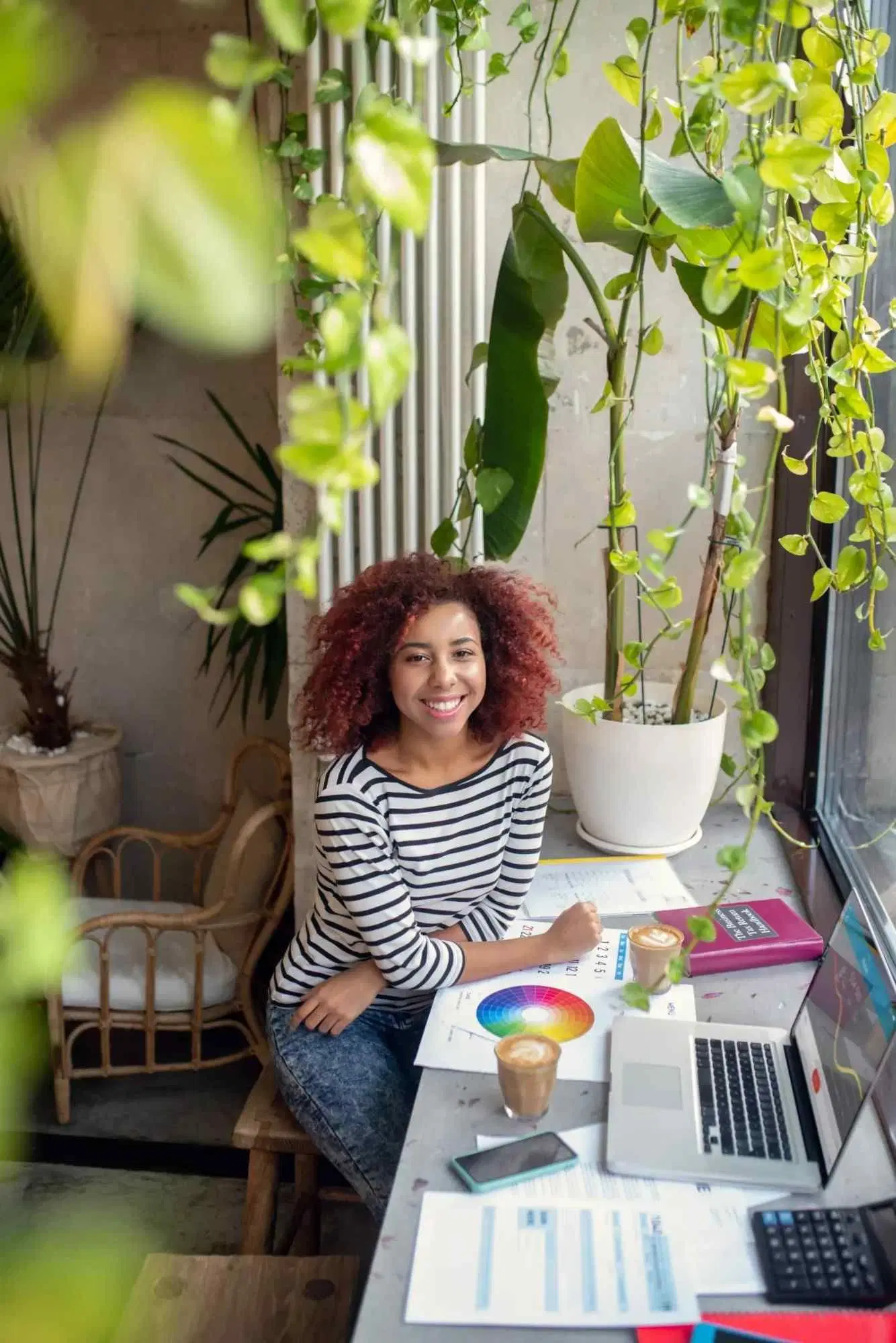 Woman working on design materials with a laptop in a plant-filled workspace.