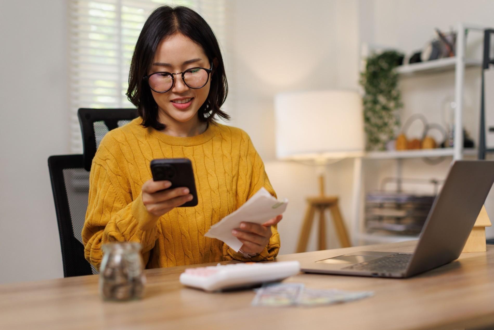 Freelancer checking her smartphone and invoices while working on a laptop from home.