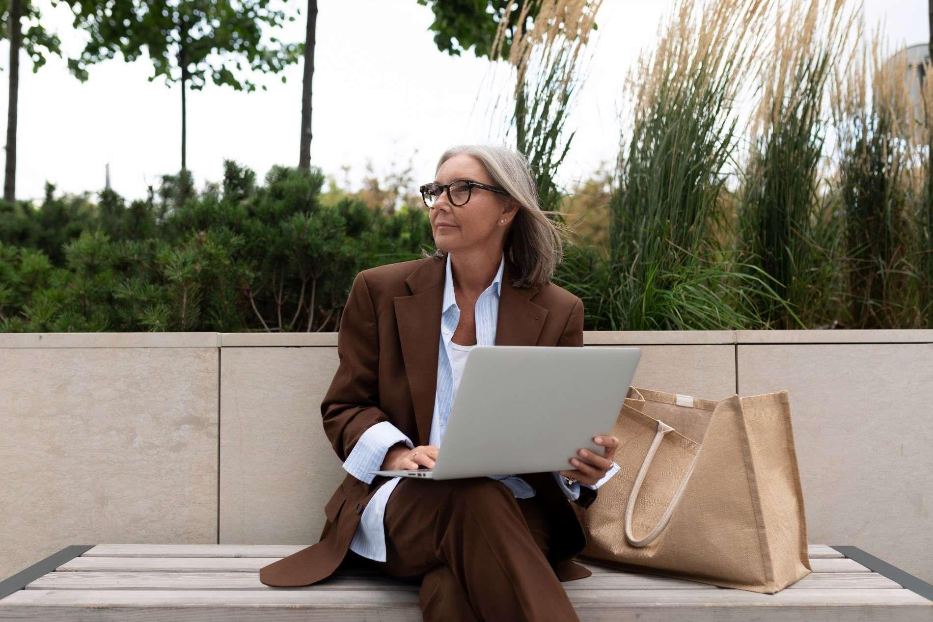 Mature woman in a brown suit working on a laptop while sitting on an outdoor bench.