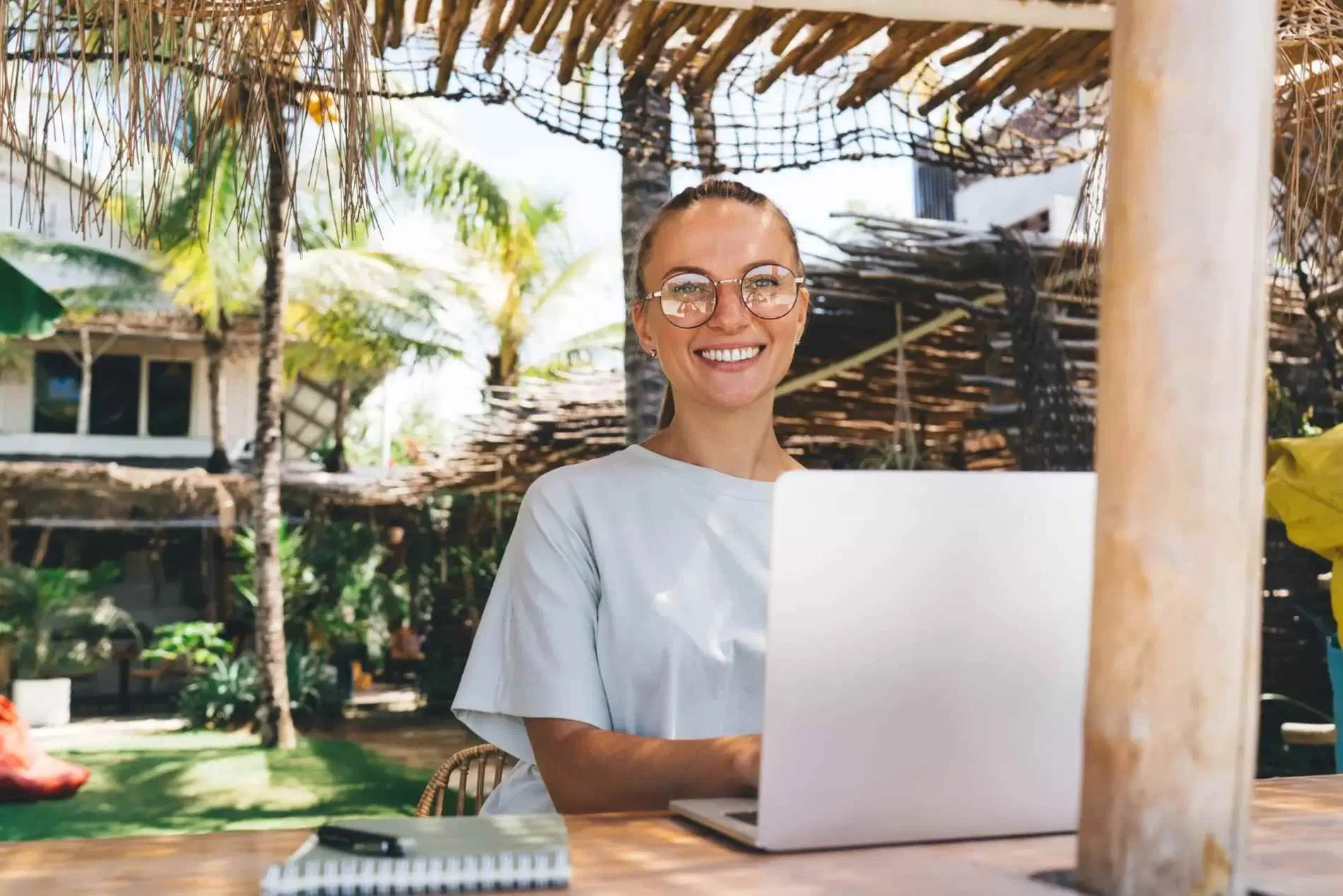 Woman smiling while working on a laptop in an outdoor tropical setting.