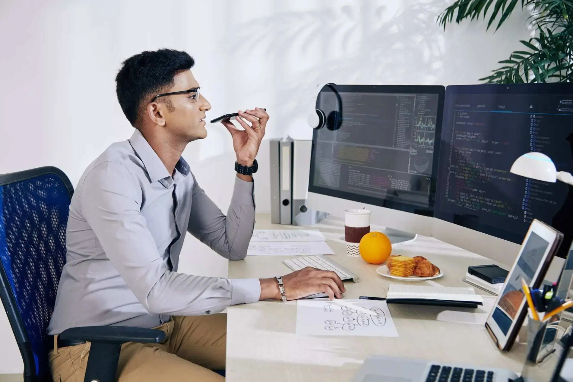 Man recording a voice message while working at a computer with code displayed on dual monitors.