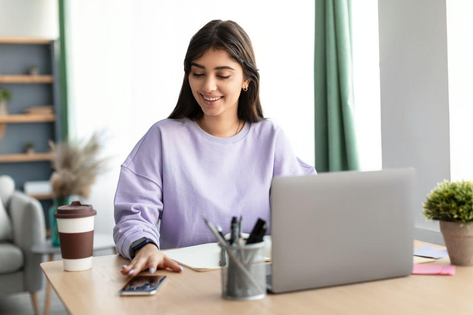 Smiling woman working on a laptop at a home desk with coffee and office supplies.