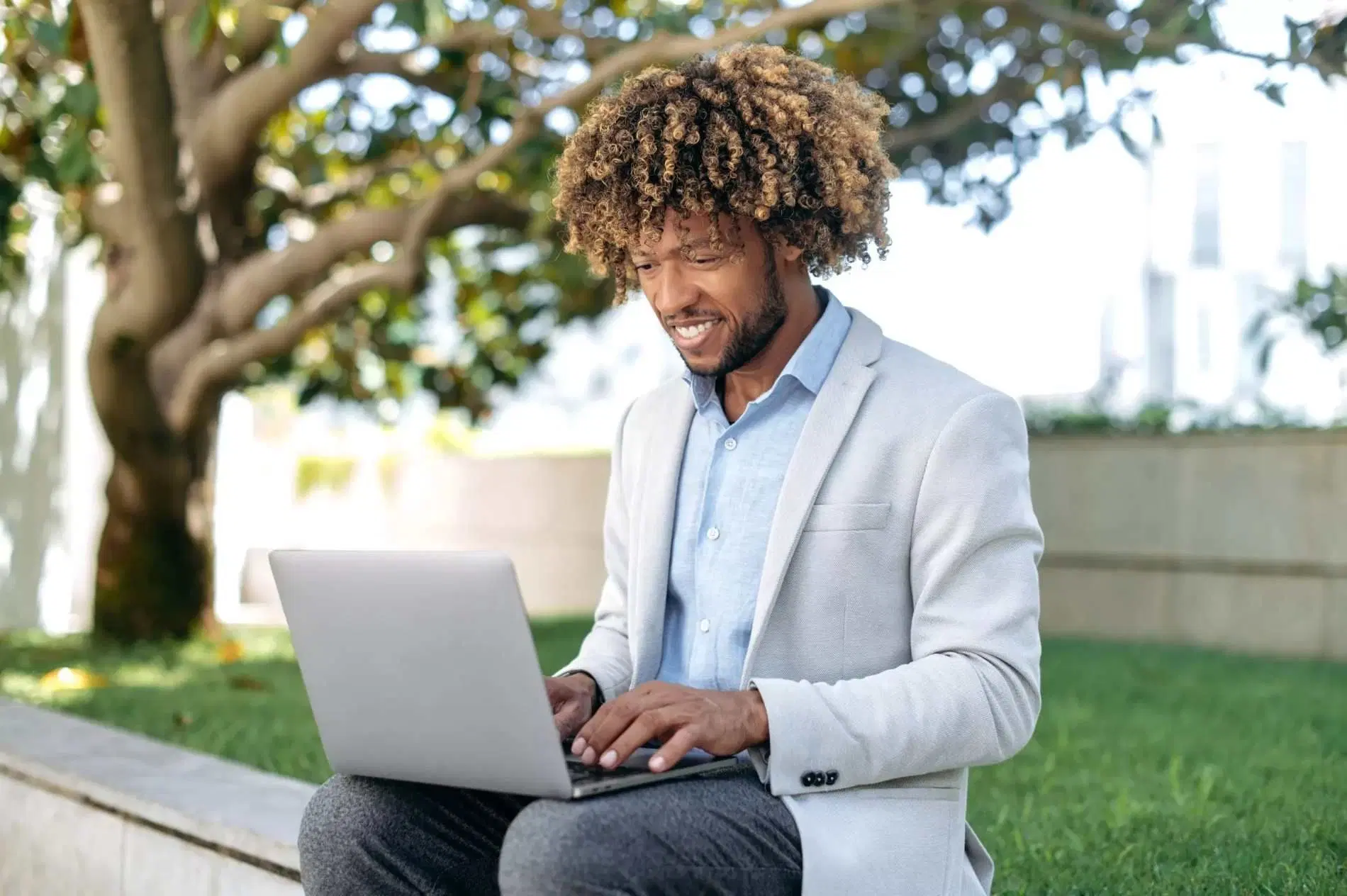 Man working on a laptop outdoors while sitting beside a tree.