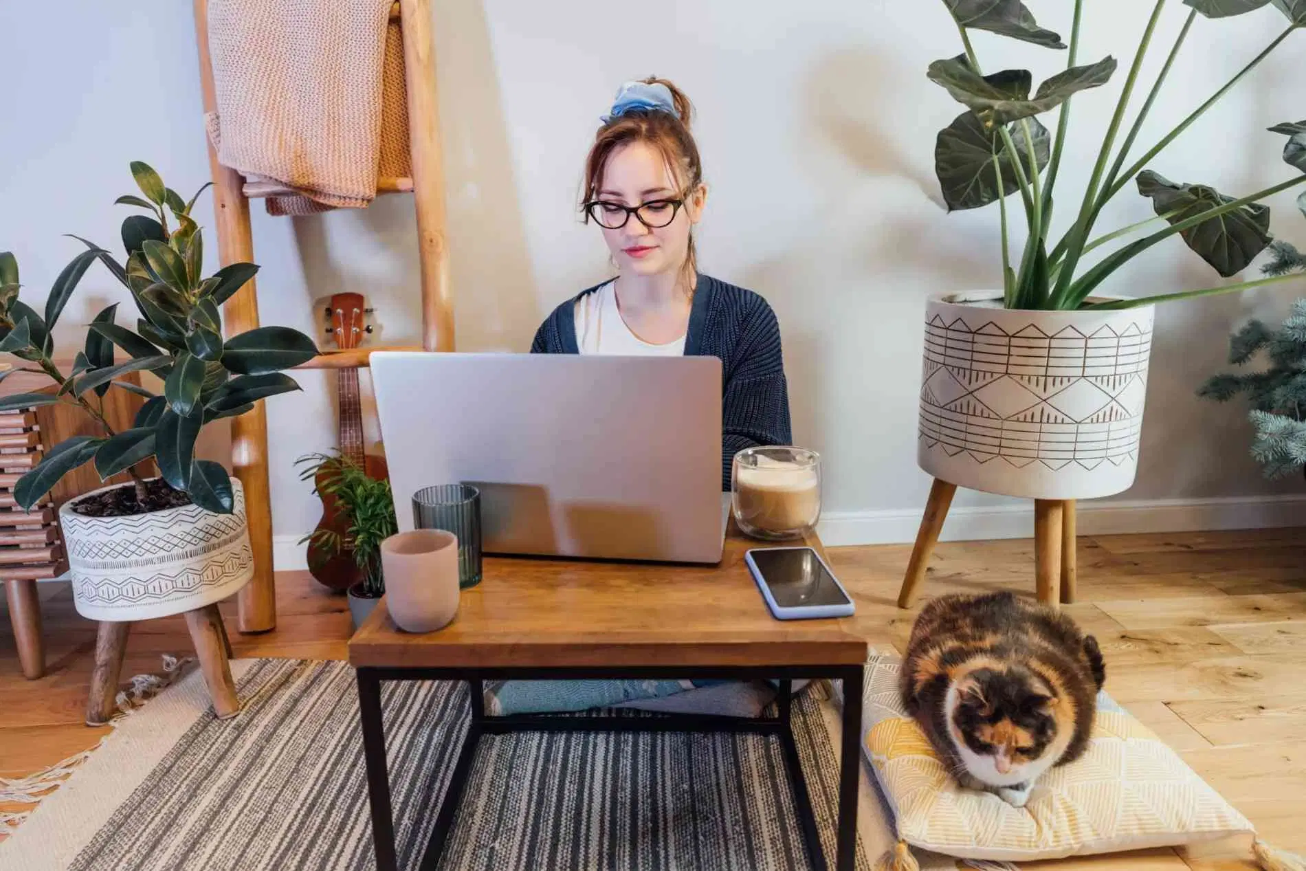 Woman working on a laptop at home with a cat beside her, surrounded by houseplants.